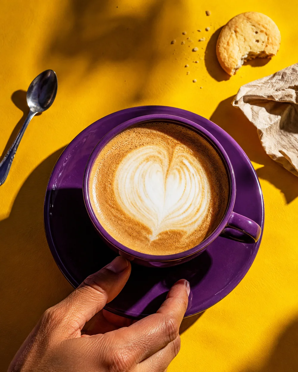 Una mano sostiene una taza morada con un corazón de espuma de leche